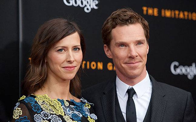 Benedict Cumberbatch in a black formal dress smiling with his wife in a blue and yellow floral dress.